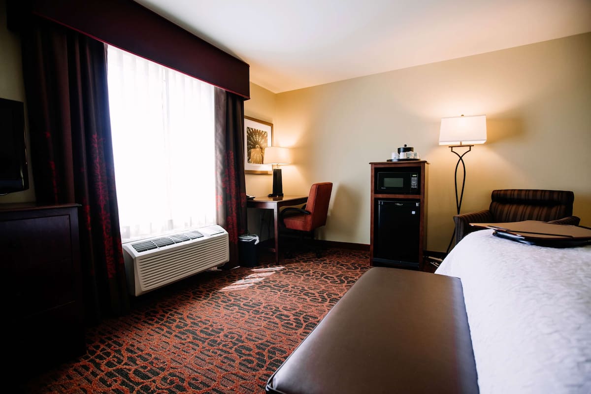 A hotel room with a bed, a desk, a chair, a TV, a lamp, and a microwave.