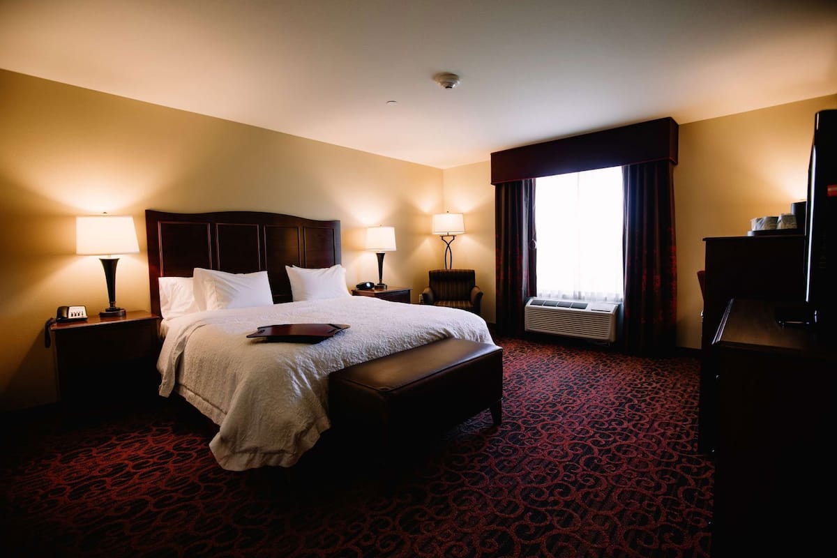 A hotel room with a large bed, two bedside tables with lamps, a chair, a desk with a computer, and a large window with curtains.
