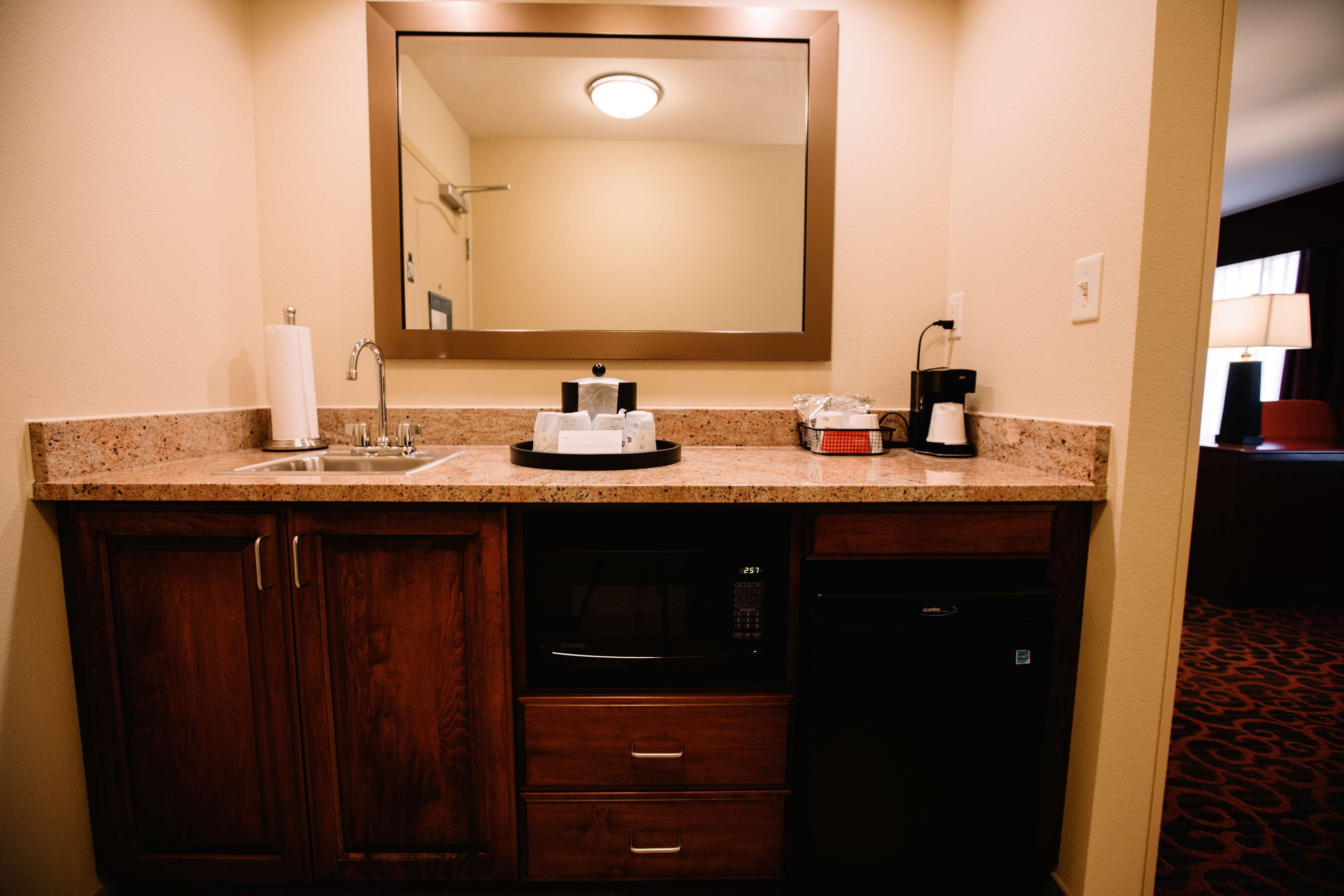 A hotel bathroom with a sink, mirror, towel dispenser, coffee maker, microwave, and a small cabinet.