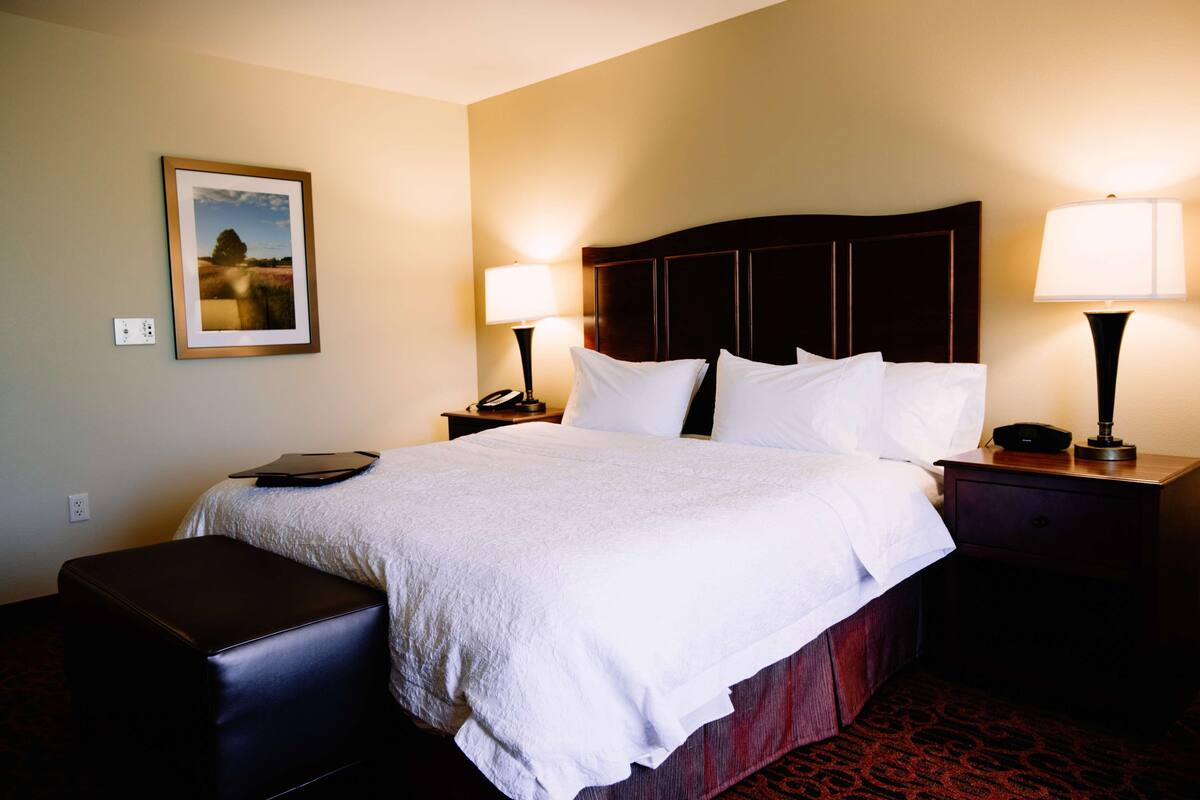 A hotel room with a large bed, two bedside tables with lamps, a nightstand, a framed picture on the wall, and a small bench.