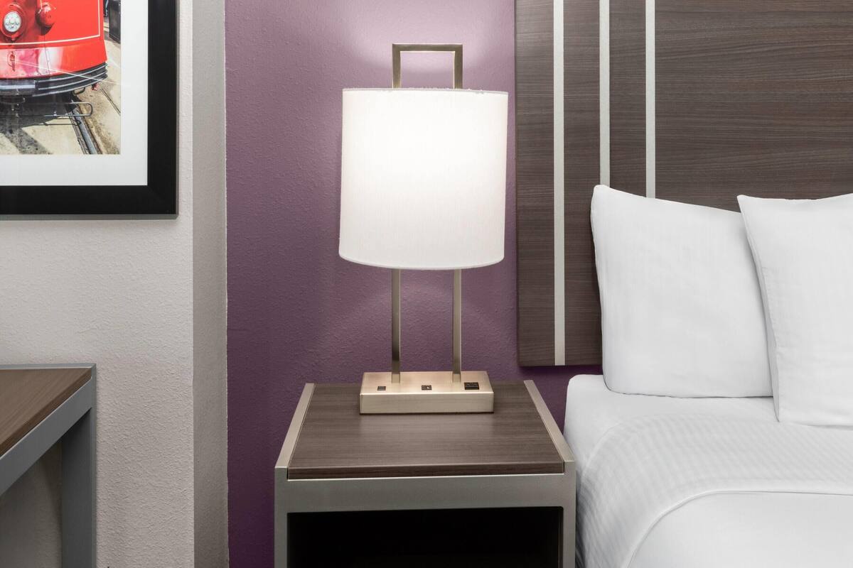 Una cama bien tendida con ropa de cama blanca, una mesita de noche con una lámpara y un cuadro en la pared.