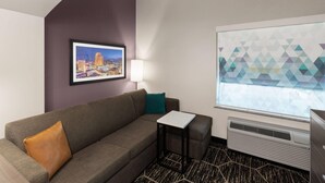 A hotel room with a gray sofa, a small table, a lamp, a framed cityscape picture, and a window with a geometric pattern. - La Quinta Inn & Suites by Wyndham Boutte (Boutte)
