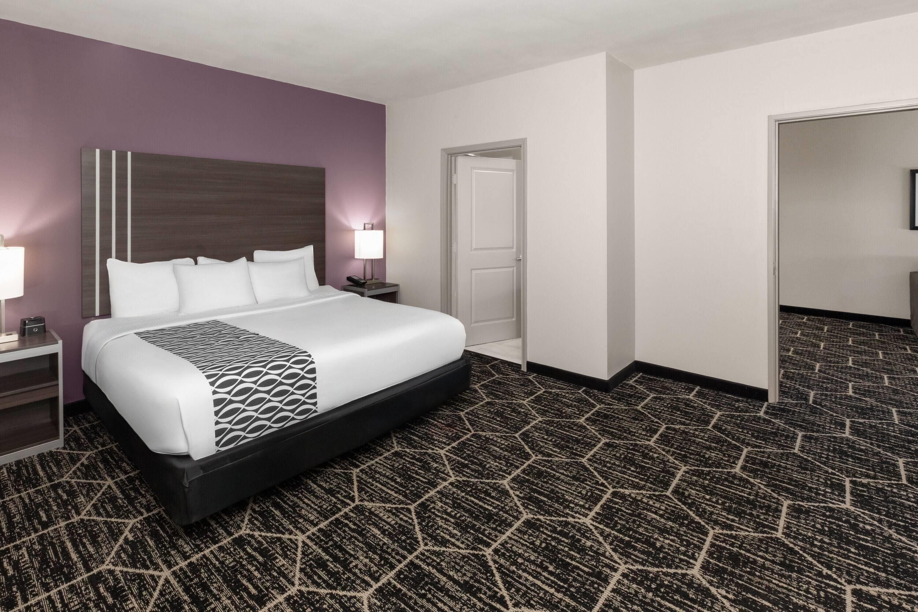 Habitación de hotel con una cama grande, dos mesitas de noche con lámparas, una alfombra estampada y una puerta que da a otra habitación.
