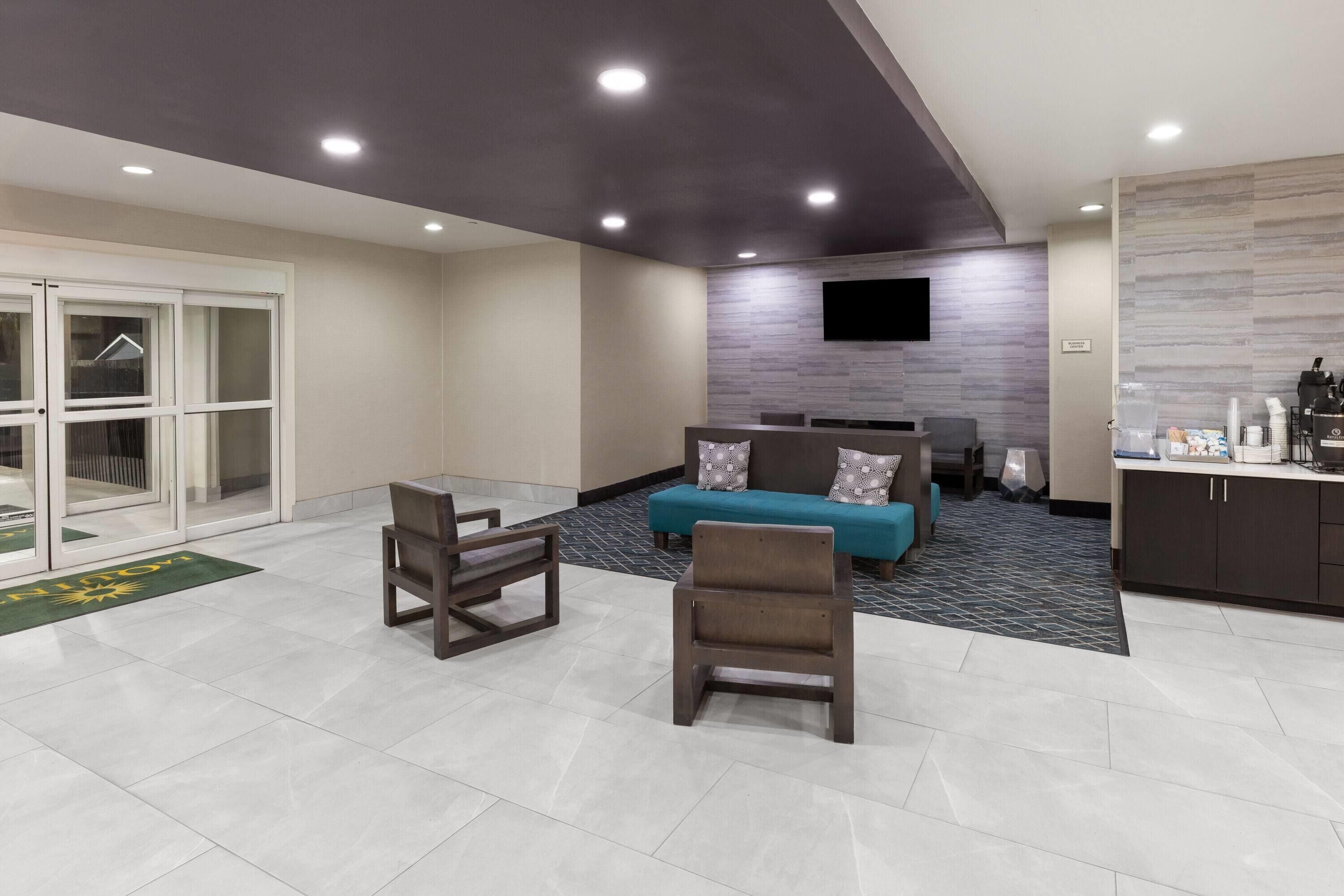 A modern hotel lobby with a sofa, chairs, a flat-screen TV, and a kitchenette area.