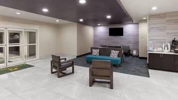 A modern hotel lobby with a sofa, chairs, a flat-screen TV, and a kitchenette area.