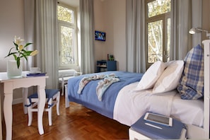 A bedroom with a bed, bedside table, chair, TV, and a window with curtains.