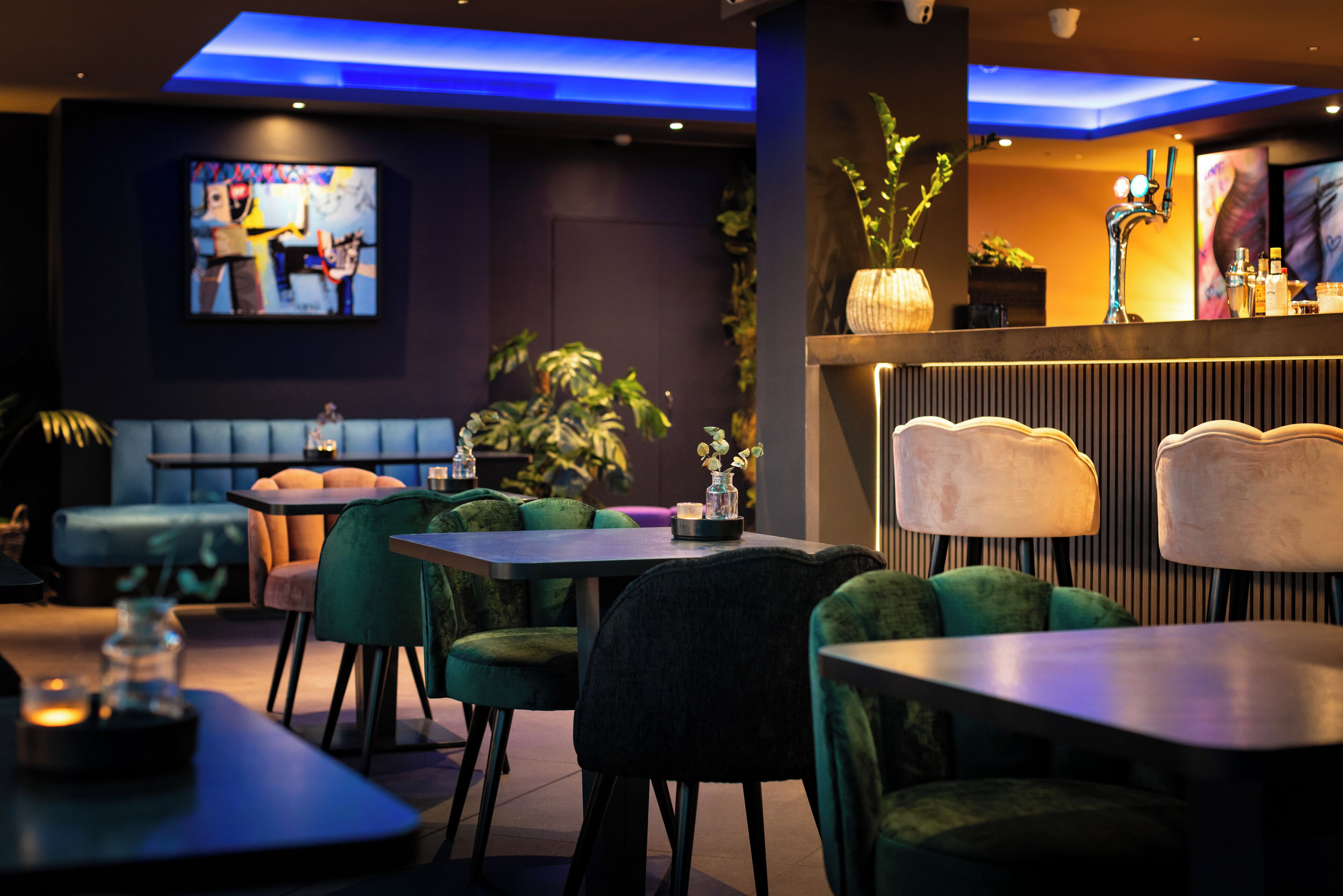 A modern bar area with green upholstered chairs, a flat-screen TV, and a bar counter with a decorative vase.