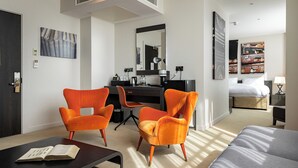 Habitación de hotel moderna con escritorio, dos sillones naranjas, una cama y obras de arte en las paredes.