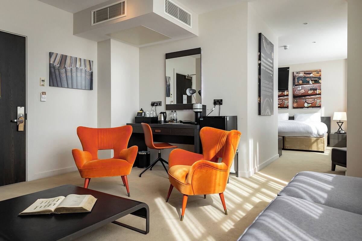 A modern hotel room with a desk, two orange armchairs, a bed, and artwork on the walls.