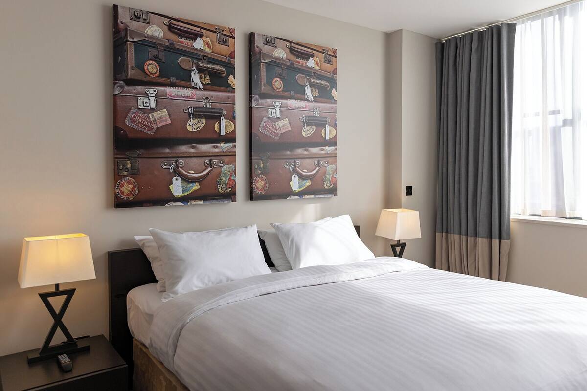 A hotel room with a bed, two nightstands, two lamps, a window with curtains, and two wall art pieces of stacked suitcases.