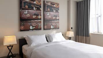 A hotel room with a bed, two nightstands, two lamps, a window with curtains, and two wall art pieces of stacked suitcases.
