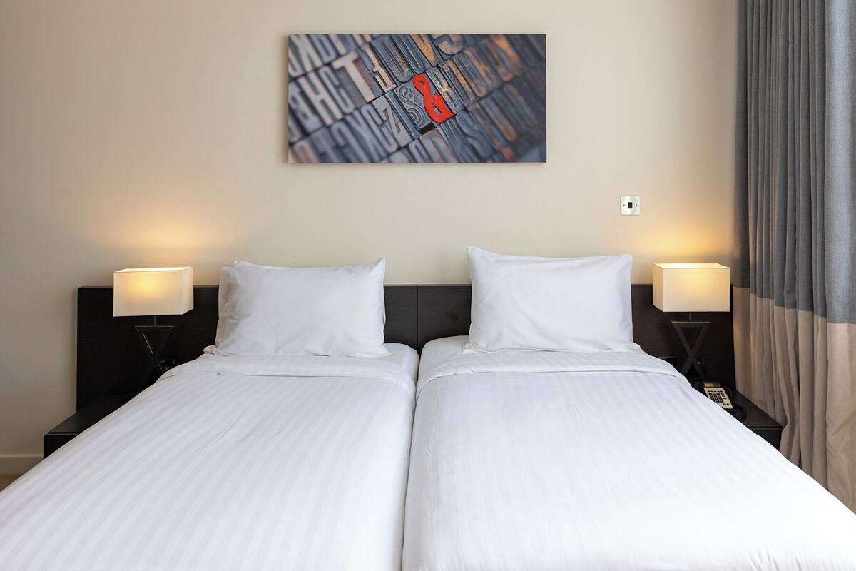 A hotel room with two single beds, a nightstand with a lamp, and a wall-mounted artwork.