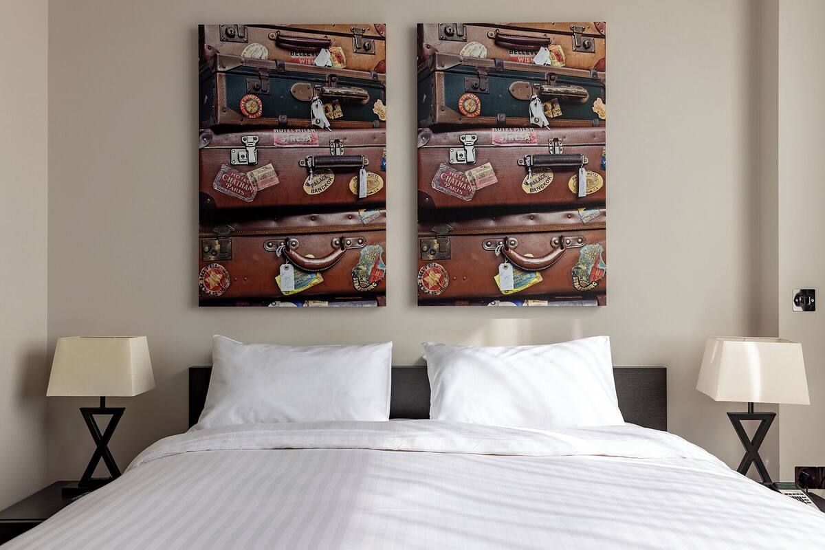 A hotel room with a bed, two bedside tables, and a wall-mounted artwork of stacked suitcases.