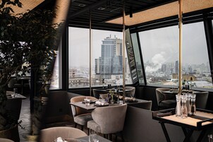 Een modern restaurant met stadszicht, tafels gedekt met glazen en bestek, en een grote potplant.