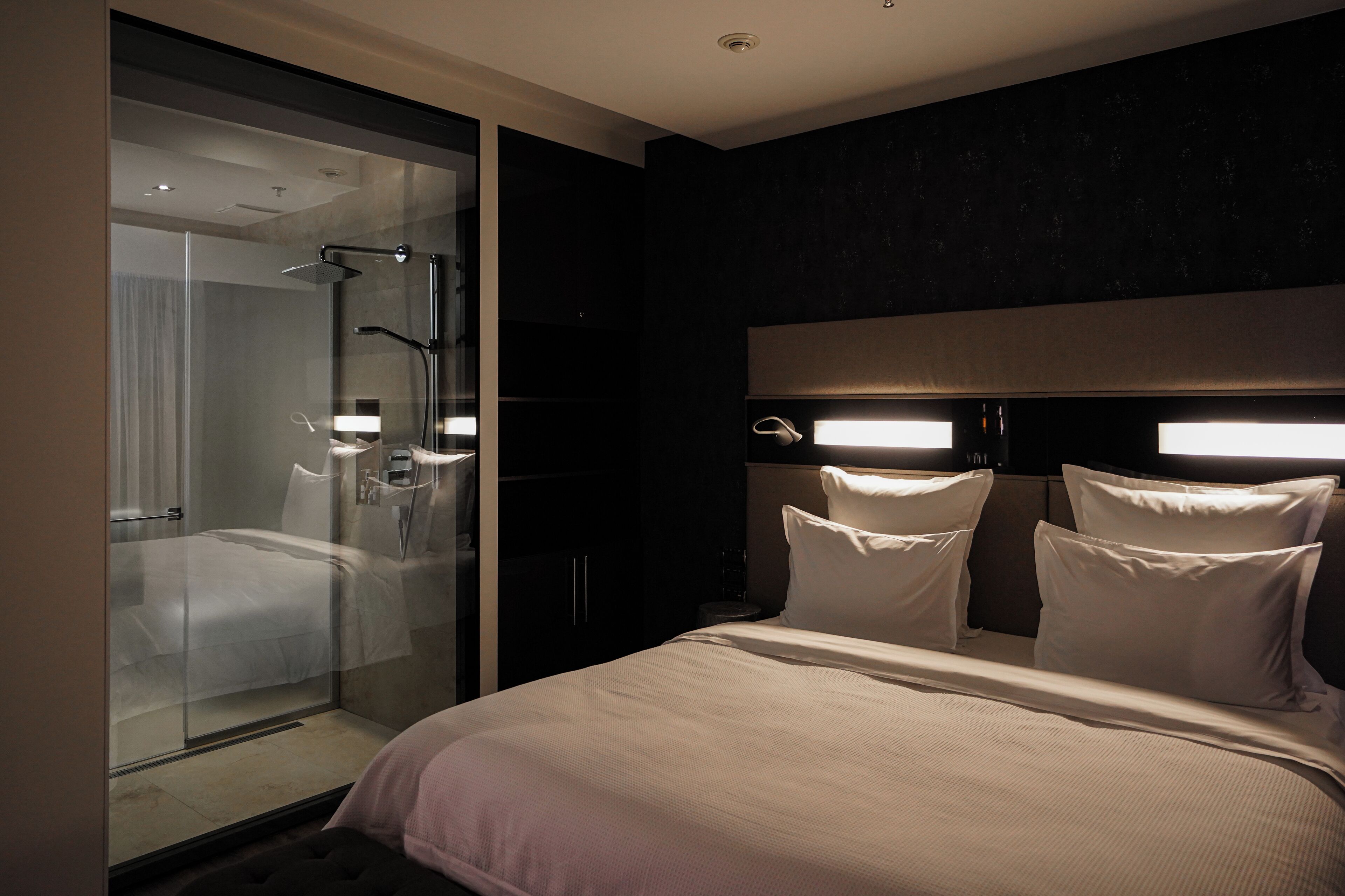 Una habitación de hotel moderna con ducha de vidrio, una cama grande con muchas almohadas y una pared de acento oscura.