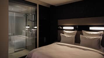 Una habitación de hotel moderna con ducha de vidrio, una cama grande con muchas almohadas y una pared de acento oscura.
