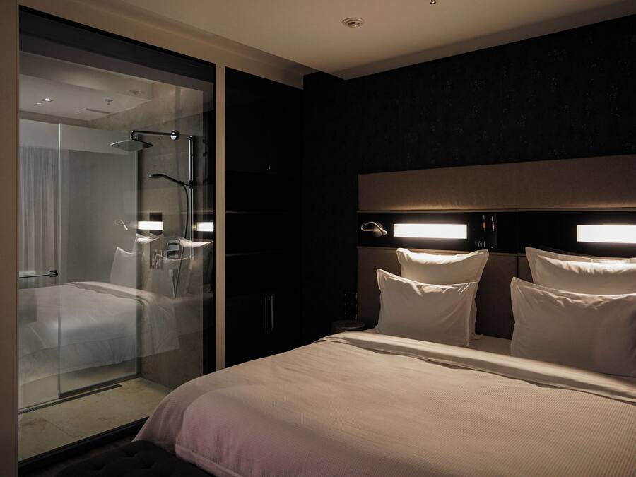 Una habitación de hotel moderna con ducha de vidrio, una cama grande con muchas almohadas y una pared de acento oscura.