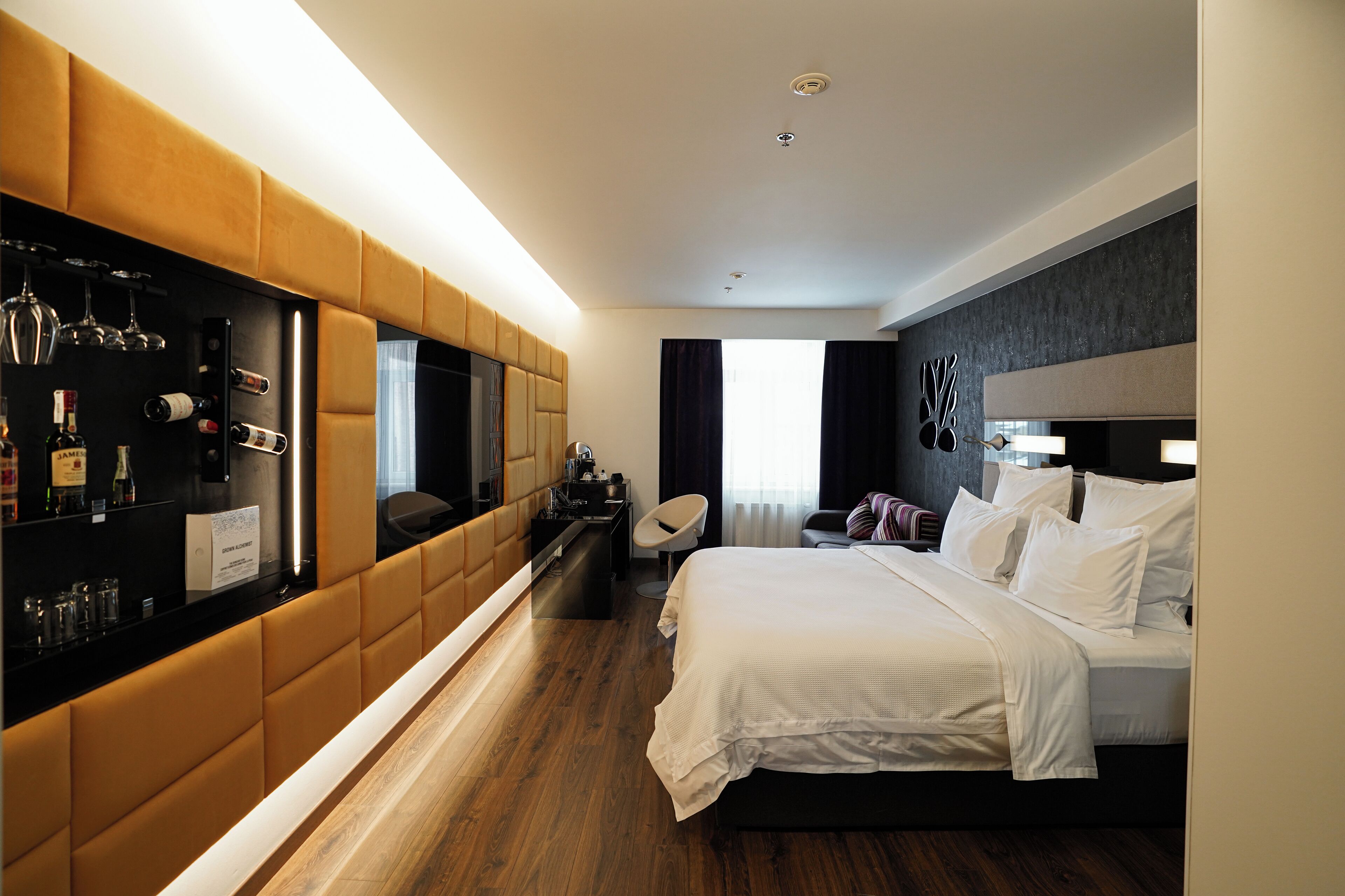 A modern hotel room with a large bed, a seating area with a sofa and armchairs, a minibar, and a TV.