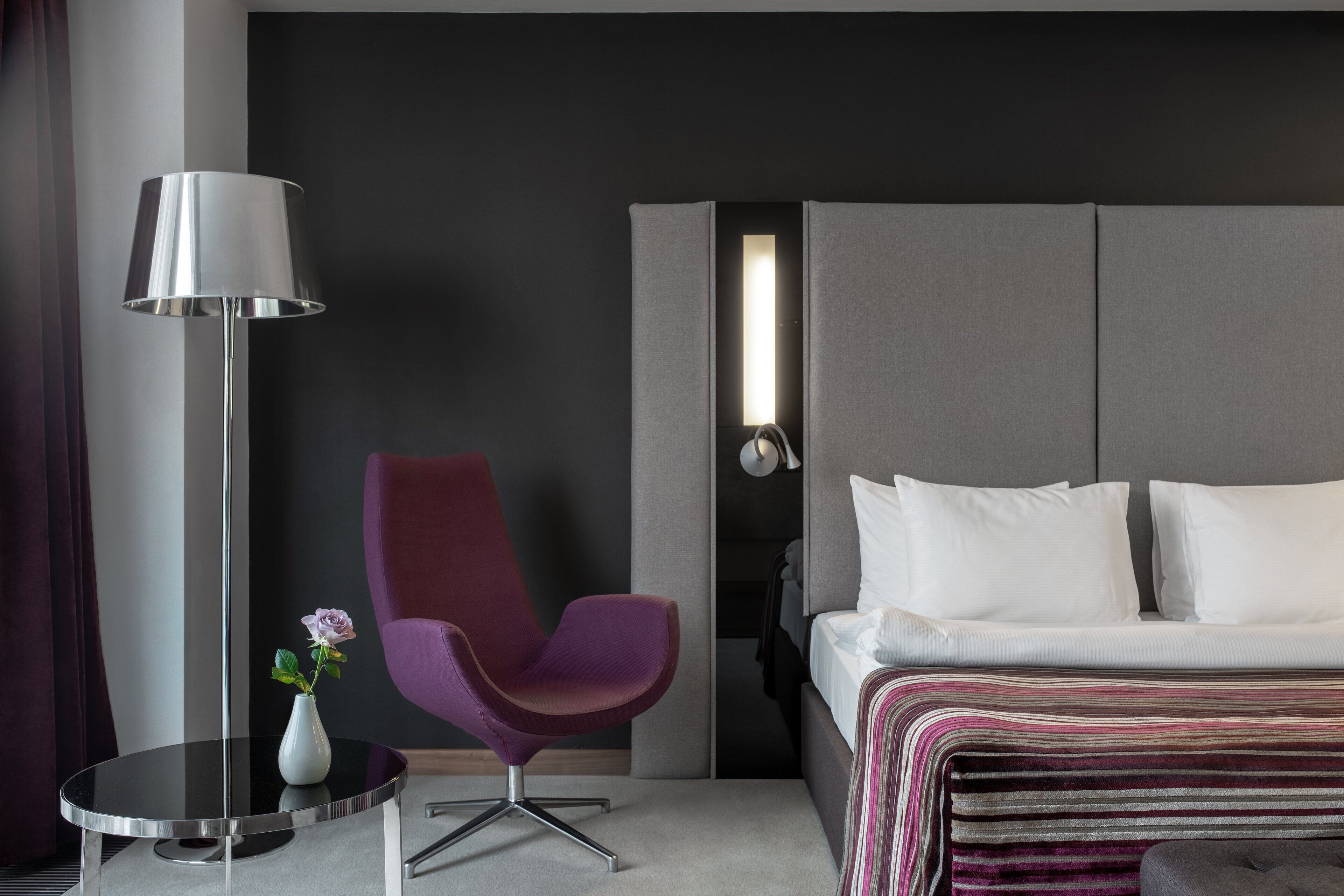 Una habitación de hotel moderna con una cama, un sillón púrpura, una mesita redonda con un florero y una lámpara de pie.