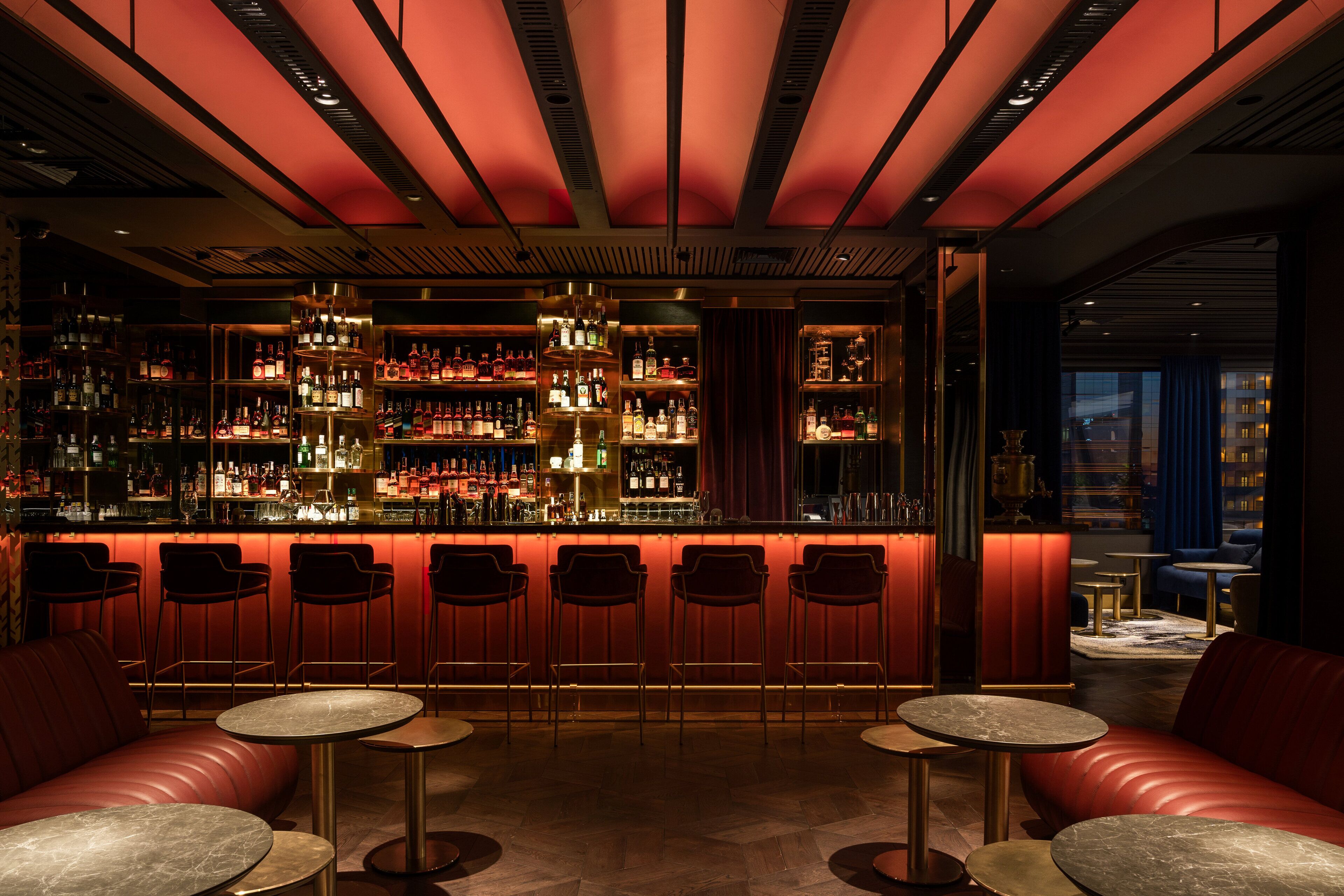 Un espace bar avec un comptoir illuminé de lumière rouge, différents types de sièges, dont un canapé rouge et des tables rondes, et une étagère bien garnie de bouteilles d’alcool.
