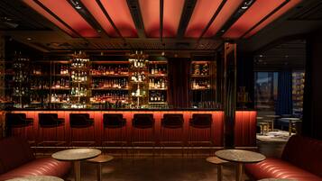 Un espace bar avec un comptoir illuminé de lumière rouge, différents types de sièges, dont un canapé rouge et des tables rondes, et une étagère bien garnie de bouteilles d’alcool.