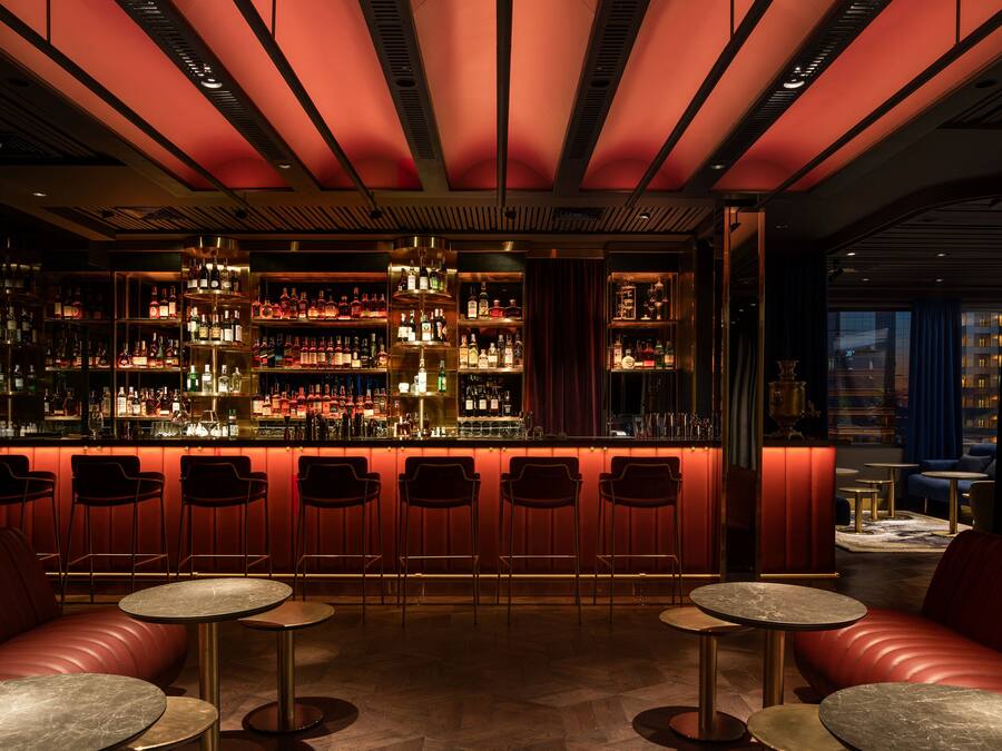 Un espace bar avec un comptoir illuminé de lumière rouge, différents types de sièges, dont un canapé rouge et des tables rondes, et une étagère bien garnie de bouteilles d’alcool.