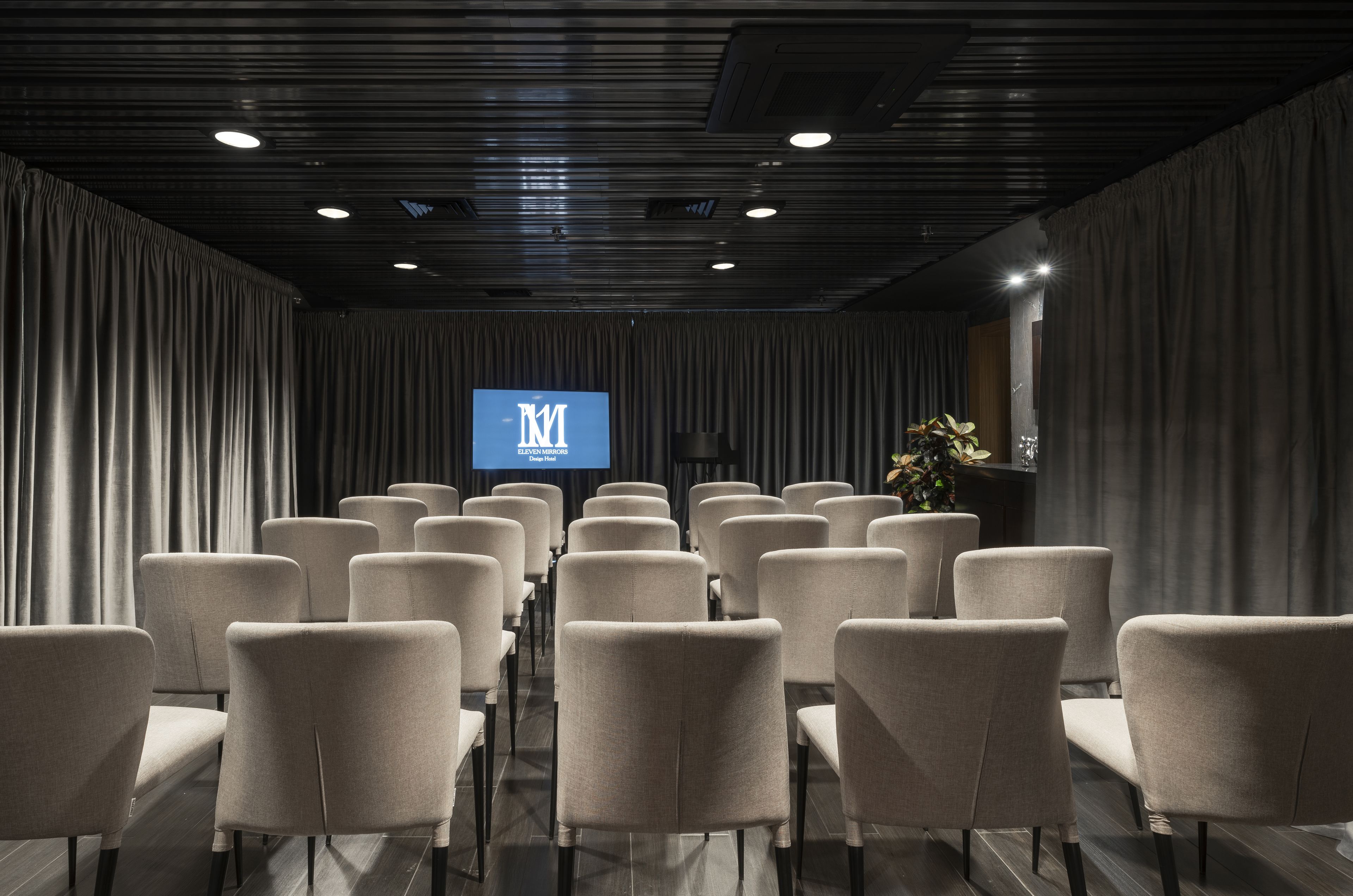 A conference room with rows of chairs, a podium, and a screen displaying the logo "M".