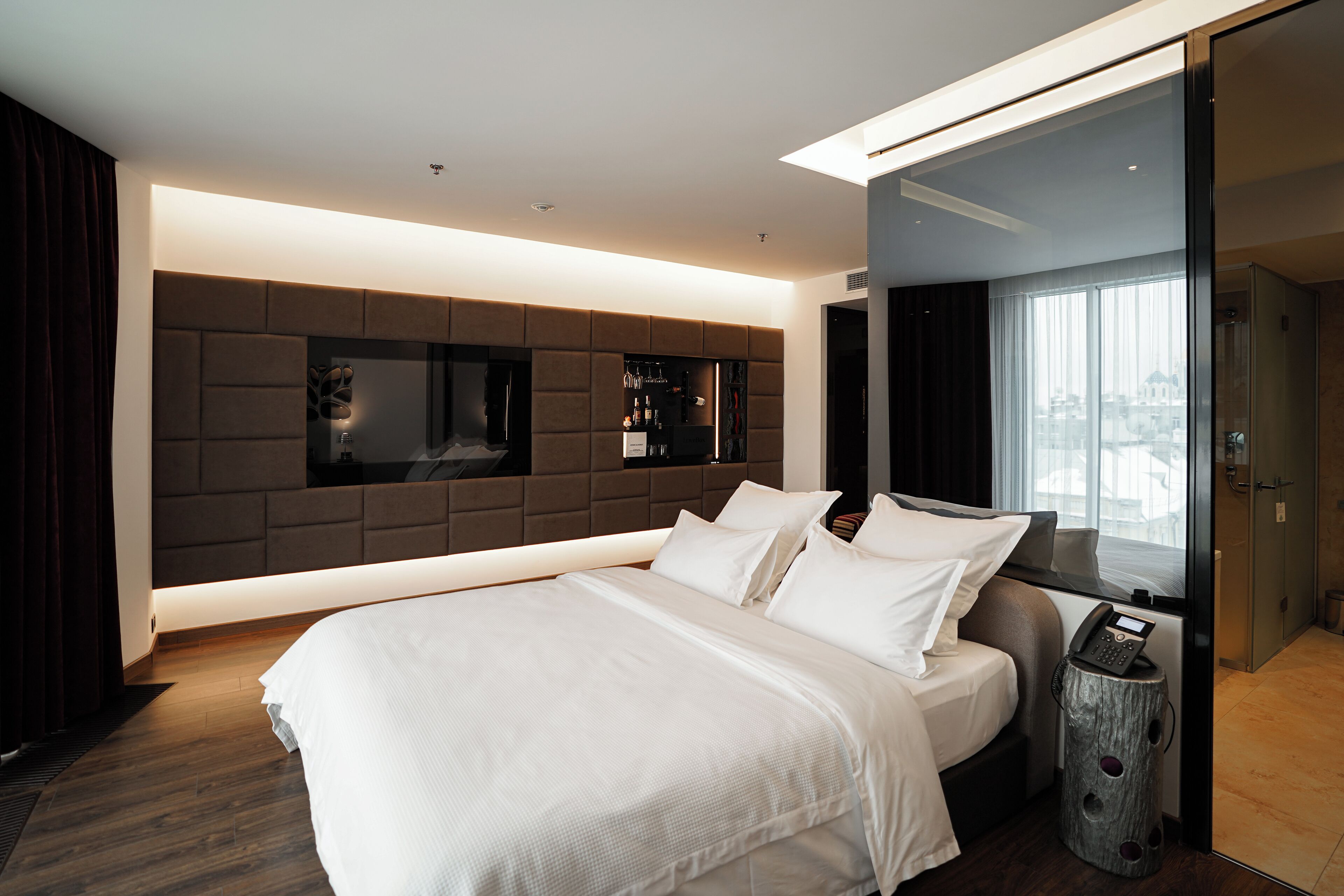A modern hotel room with a large bed, a built-in TV, a minibar, and a view of the city through the window.