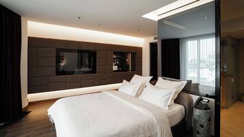 A modern hotel room with a large bed, a built-in TV, a minibar, and a view of the city through the window.