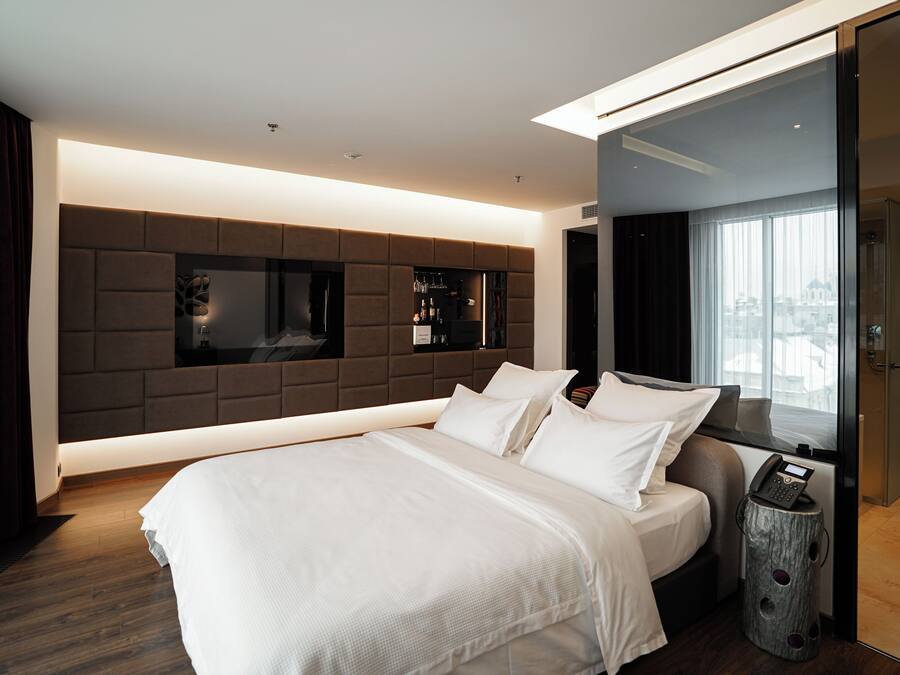A modern hotel room with a large bed, a built-in TV, a minibar, and a view of the city through the window.