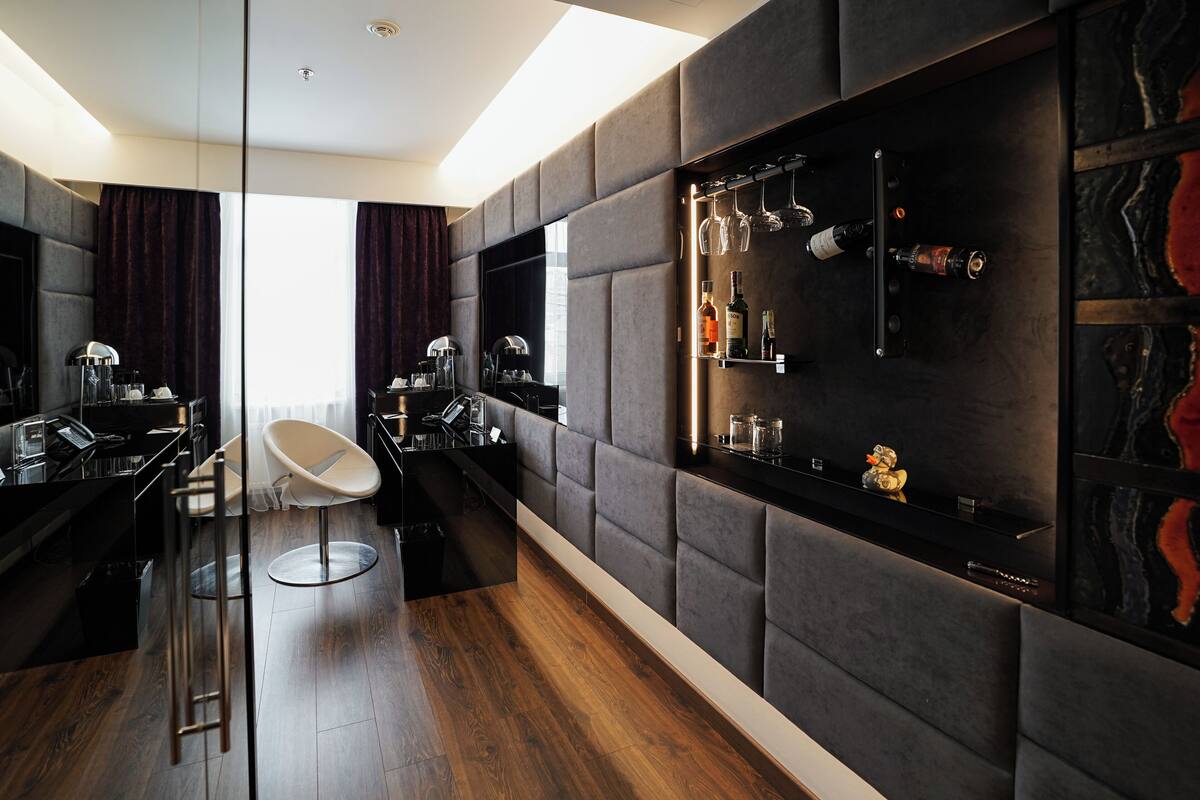 A modern hotel room with a dark wood floor, a glass partition, a sofa, a dining area, and a wall-mounted wine rack.