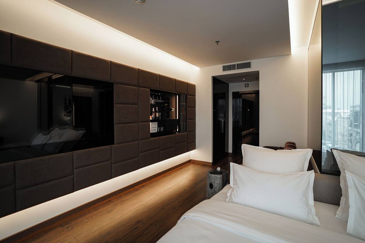 A modern hotel room with a large bed, a built-in TV, a minibar, and a city view.