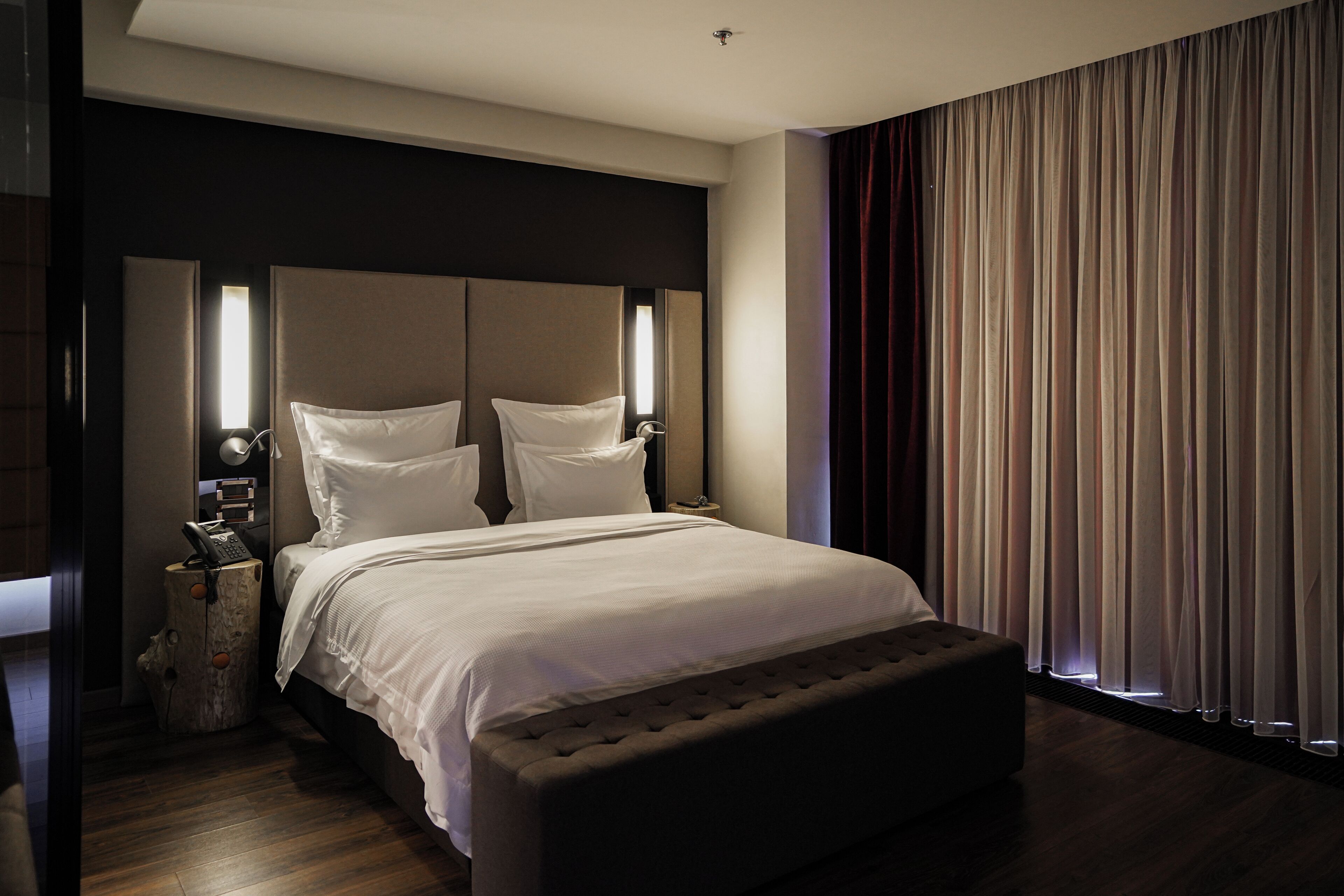 Habitación de hotel moderna con una cama grande, una mesita de noche con lámpara y vistas de la ciudad a través de cortinas.