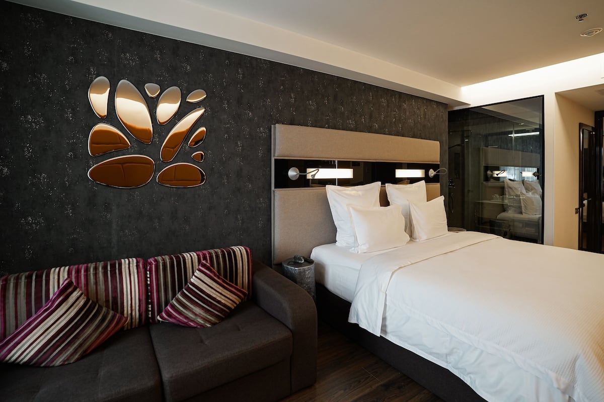A modern hotel room with a large bed, a sofa with striped cushions, a wall-mounted light fixture, and a glass partition.