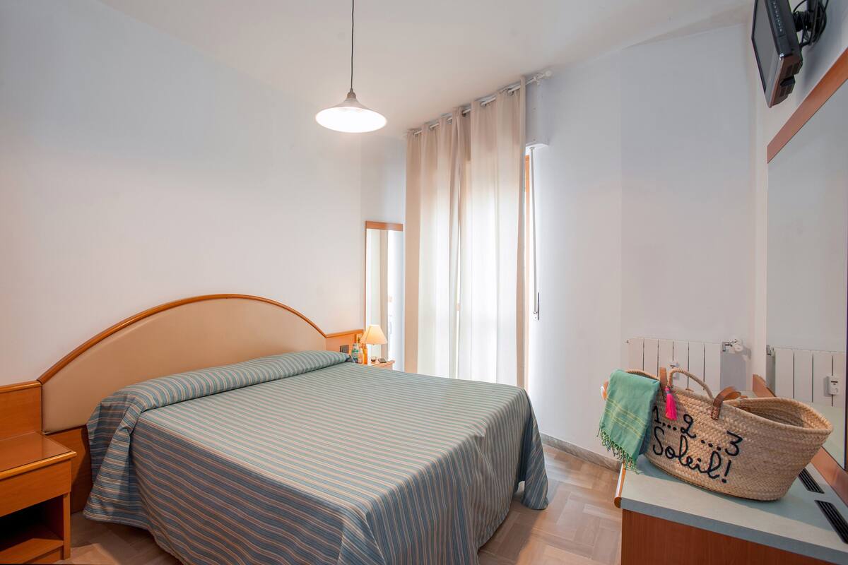 Une chambre à coucher comprenant un lit, une table de chevet, une lampe, une fenêtre avec des rideaux et un radiateur.