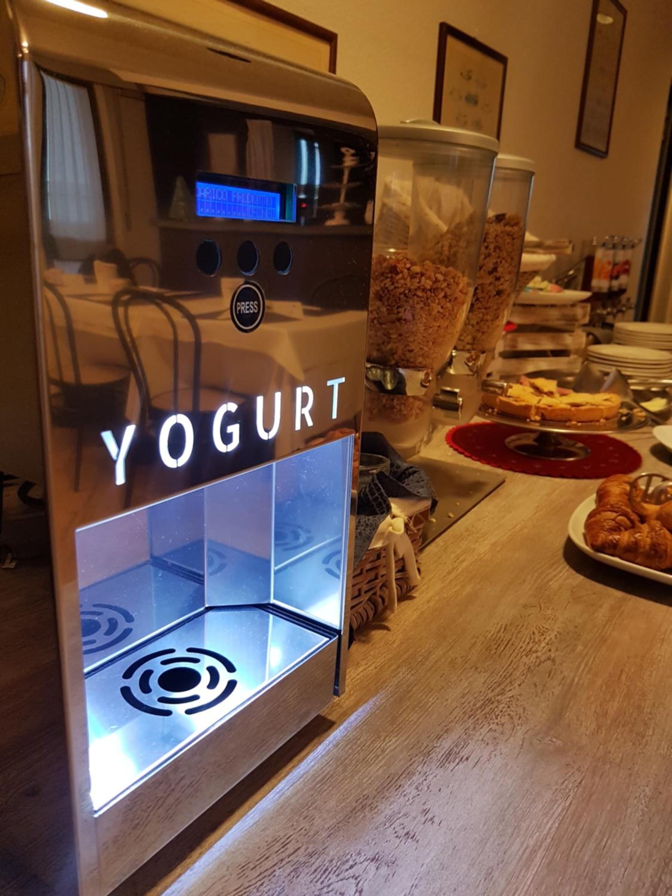 Una macchina per yogurt con display digitale e pulsante, posizionata su un tavolo di legno con cibo e un sottopiattino rosso nelle vicinanze.