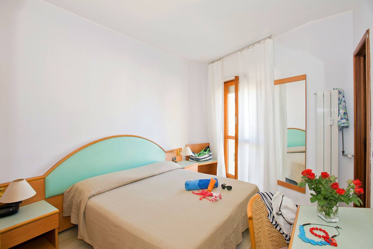 Une chambre d’hôtel avec un lit, une table de chevet, une lampe et une petite table sur laquelle est posé un vase de fleurs.