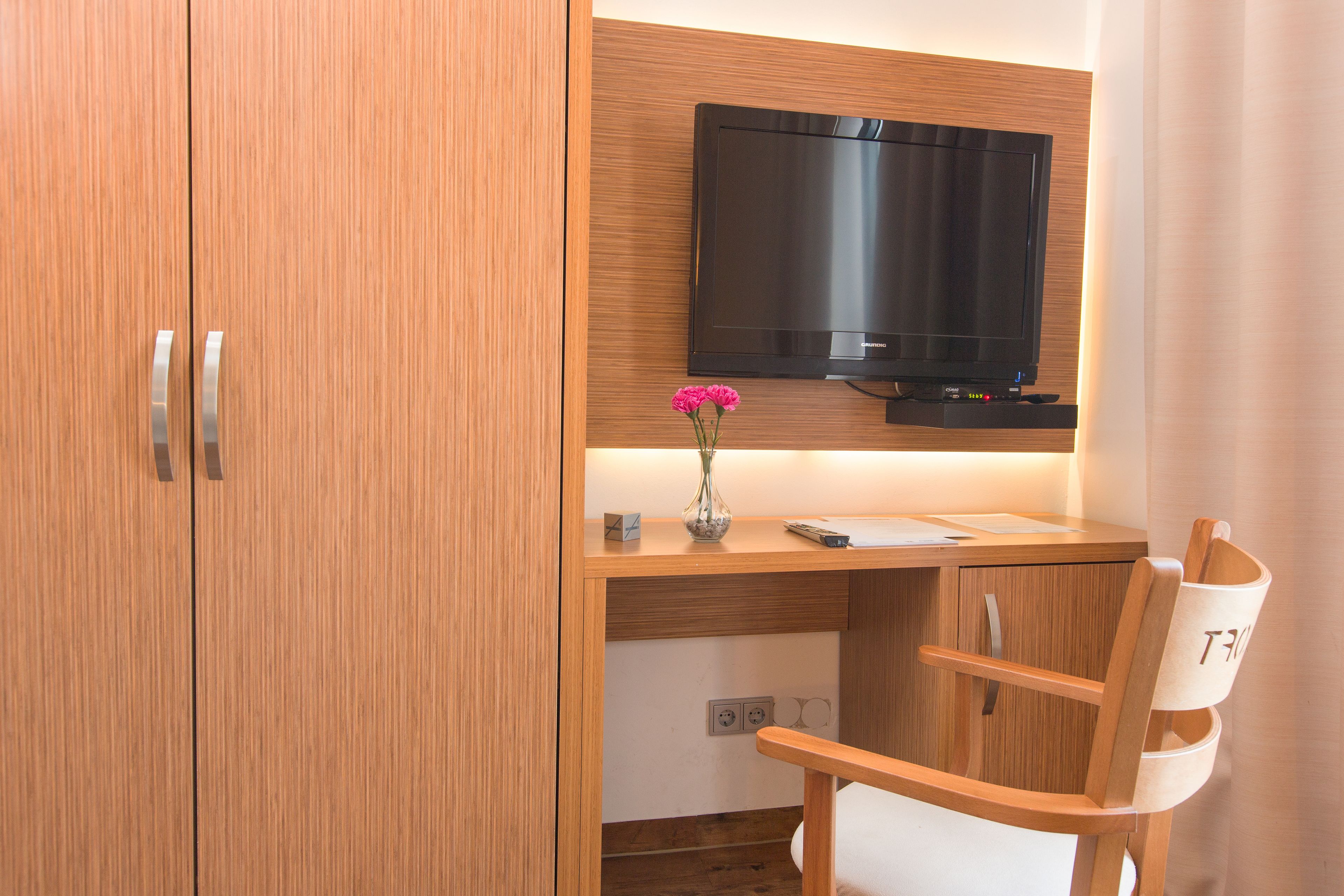 Ein modernes Zimmer mit einem Holzbüro, einem flachbildfernseher darüber montiert, einer Vase mit Blumen und einem Sessel mit einem Logo.