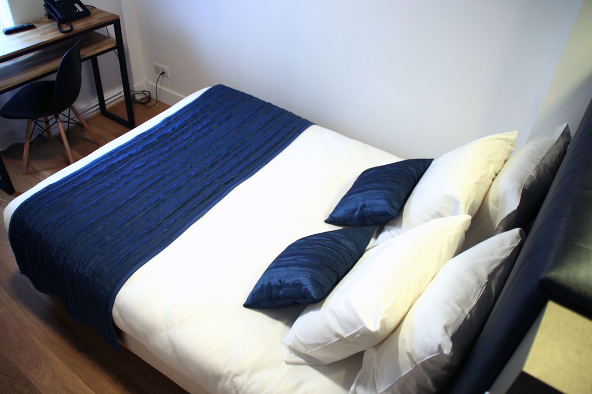 A neatly made bed with a blue and white bedspread, two pillows, and a wooden desk with a chair and a telephone.
