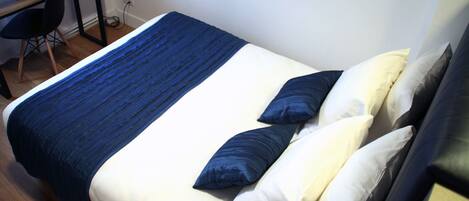 A neatly made bed with a blue and white bedspread, two pillows, and a wooden desk with a chair and a telephone.