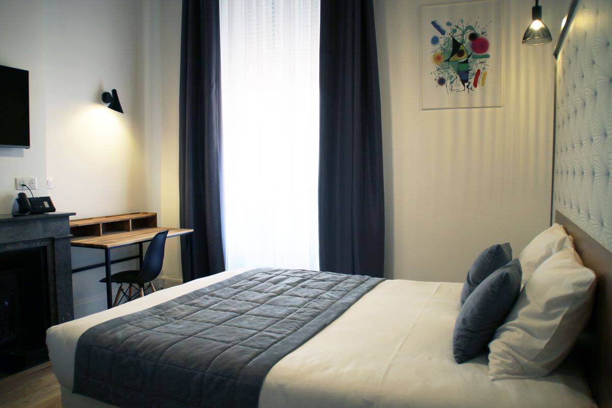 Habitación de hotel con una cama, un escritorio y una silla. Se ve una ventana con cortinas, un cuadro en la pared y una lámpara.