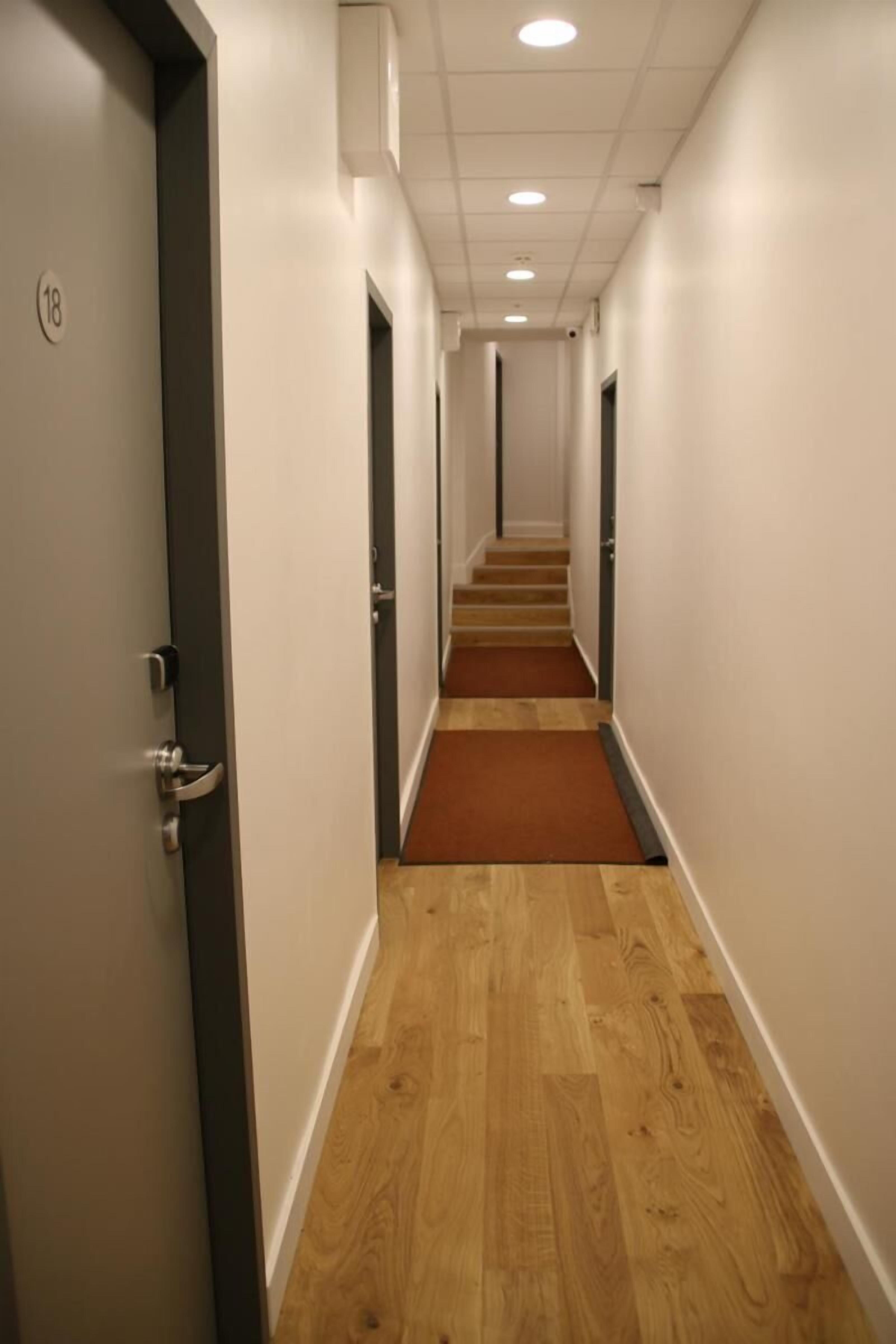 Un pasillo de hotel con piso de madera, paredes blancas y una alfombra marrón que conduce a una escalera.