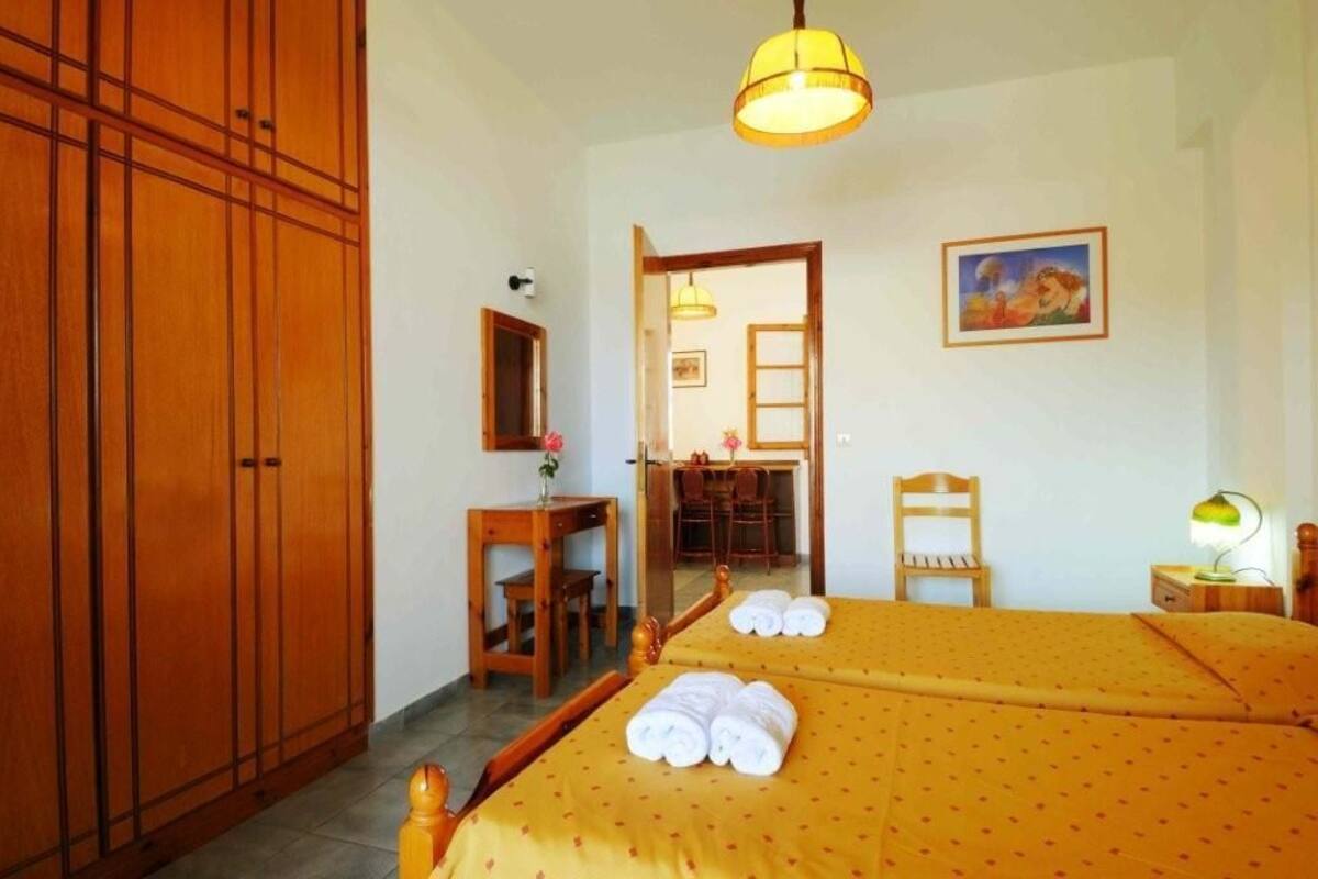 Habitación de hotel con dos camas, un escritorio, una silla, un armario y un área de comedor visible a través de una puerta abierta.
