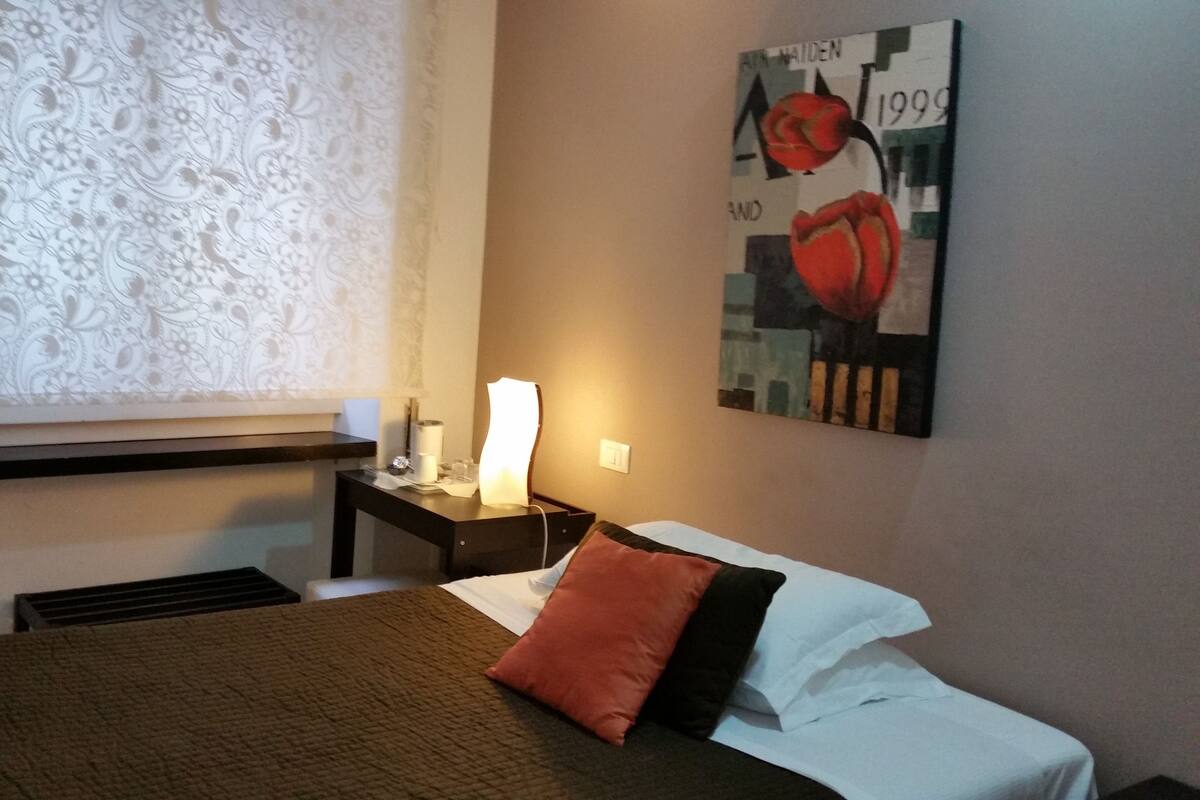 Camera d'albergo con un letto, un comodino, una lampada, una finestra con tende a motivi e un quadro appeso al muro.