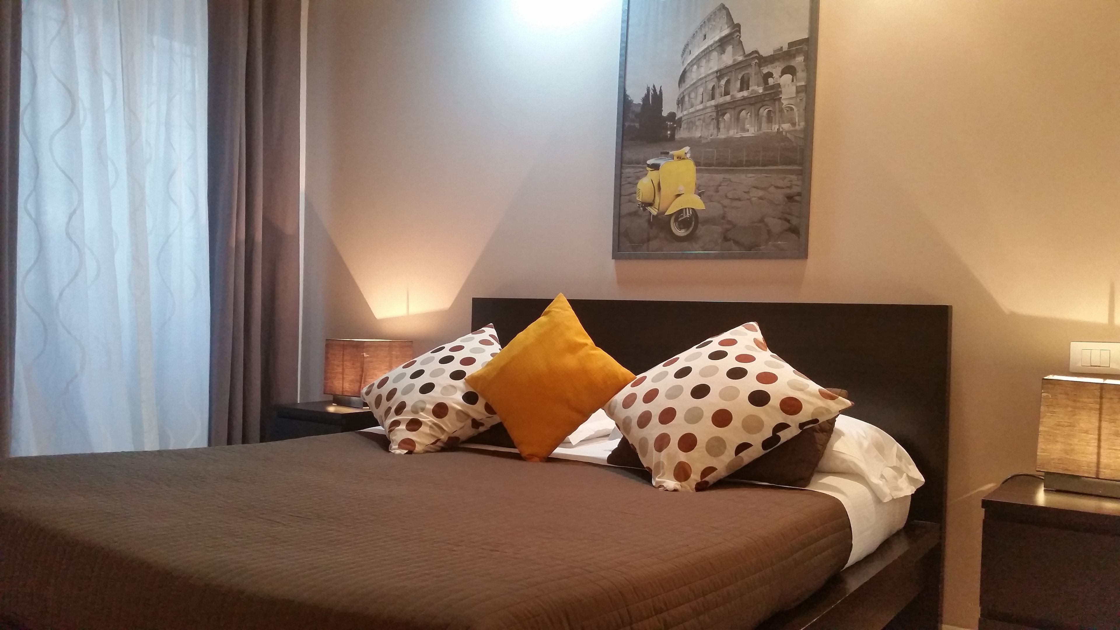 A hotel room with a neatly made bed, a nightstand with a lamp, and a wall-mounted picture of the Colosseum.