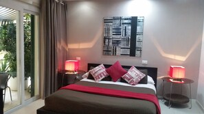 A bedroom with a bed, two bedside tables with lamps, a large abstract painting on the wall, and a view of greenery through a sliding door. - InnVatican (Rome)