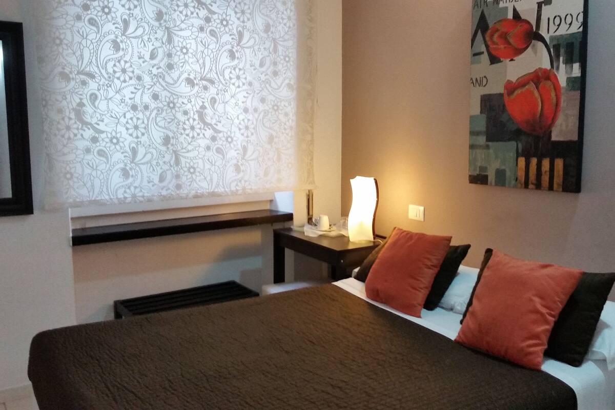 Una camera d'albergo con un letto, un comodino con una lampada, una finestra con tende a motivi e un quadro appeso al muro.