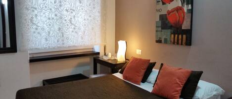 A hotel room with a bed, a nightstand with a lamp, a window with patterned curtains, and a painting on the wall.