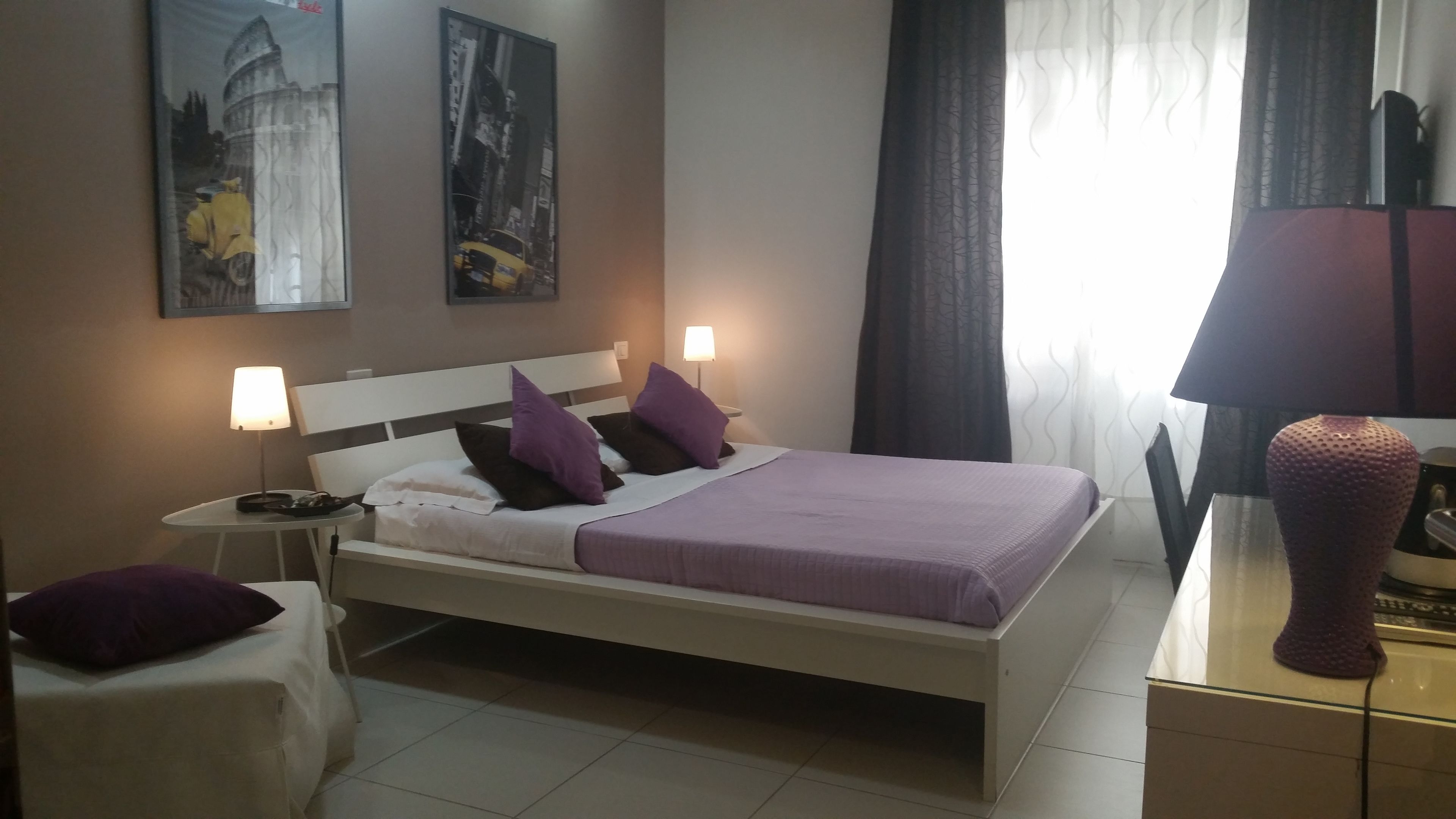 Habitación de hotel con cama, dos lámparas de noche, una mesita de noche, una silla, un jarrón grande y dos cuadros en la pared.