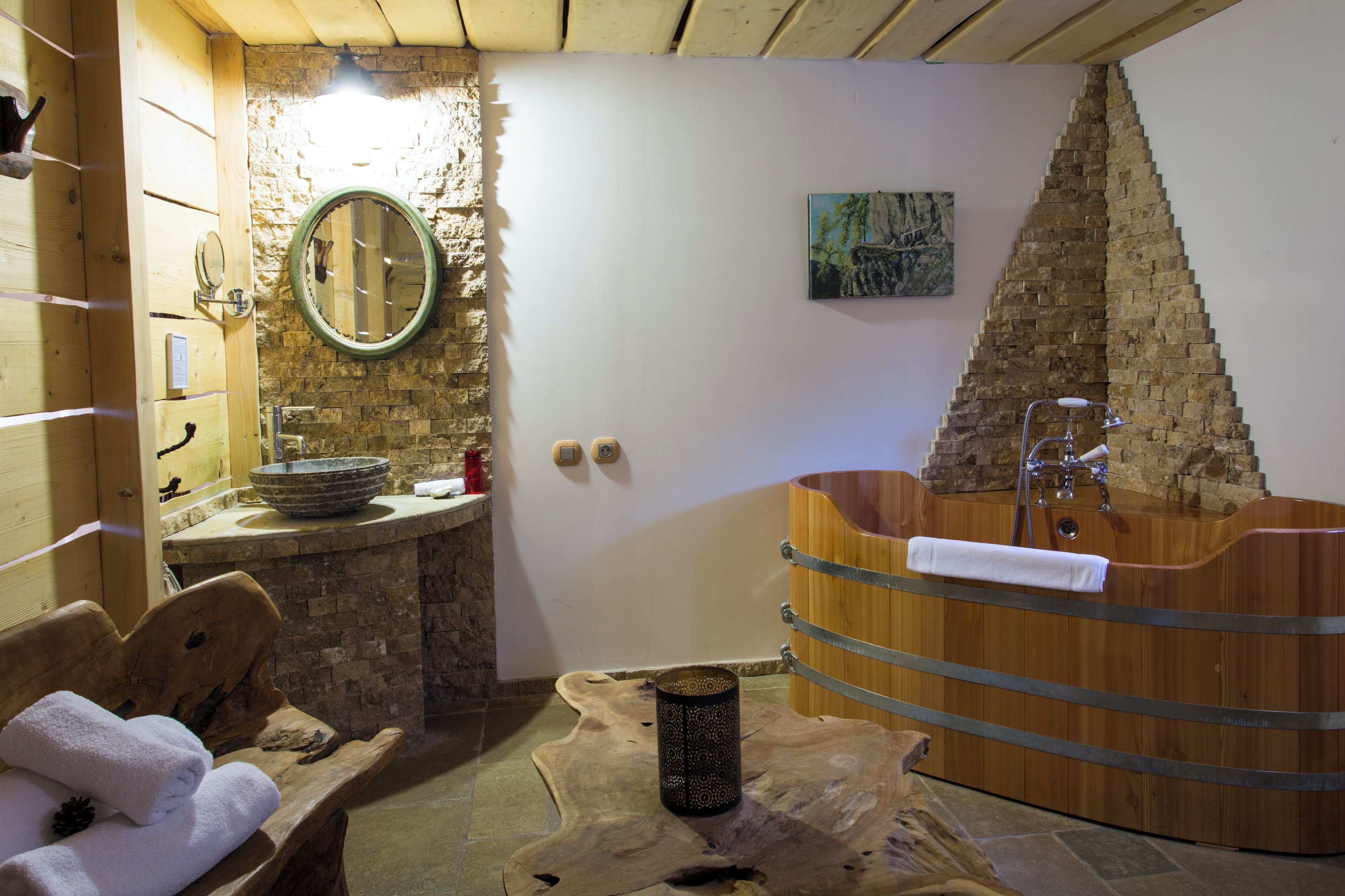 Una spa con stanza in legno, parete in pietra, vasca da bagno in legno, panchetta con asciugamani e specchio tondo.